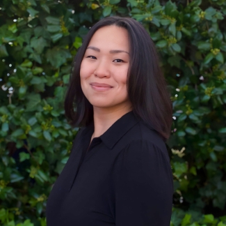 A woman with dark, shoulder-length hair smiling in a professional headshot against a lush green ivy background.