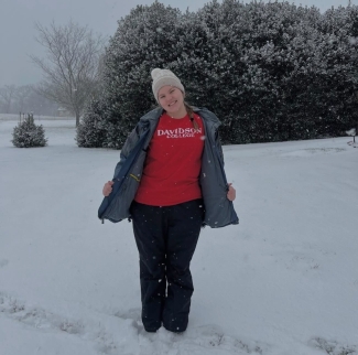 A smiling person stands in a snowy field wearing a red Davidson College shirt, a grey winter hat, and a dark open jacket.