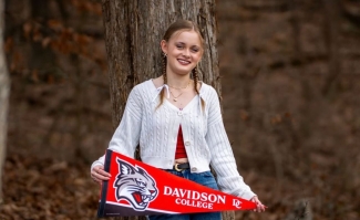A person with braided hair leans against a tree trunk in a wooded area, smiling and holding a red and black Davidson College pennant featuring a wildcat logo.