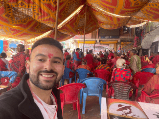 A smiling man with a red tilak on his forehead takes a selfie in front of a large outdoor gathering seated under a vibrant, patterned orange and yellow canopy.