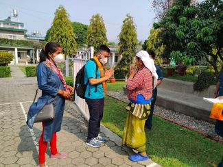 A man wearing a backpack and an orange garland exchanges a traditional Namaste greeting with an elderly woman in patterned clothing on a paved walkway in Nepal.