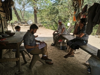 A man sits at a rustic wooden table under a shaded outdoor structure, writing in a notebook while two older adults and a child sit nearby in a rural, wooded setting.