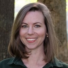 A smiling woman with shoulder-length brown hair wearing a dark green collared shirt, pictured against a blurred background of trees.