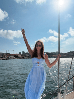 A smiling woman in a blue and white striped dress and sunglasses poses on a sailboat with a coastal city and cable cars in the background.