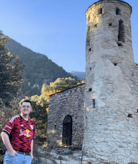 A smiling man in a red and black patterned jersey stands before an ancient, cylindrical stone tower and a matching stone building set against a backdrop of forested mountains and a clear blue sky.
