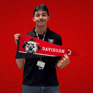 A person in a black polo shirt and lanyard stands against a red textured background, holding a red Davidson College pennant with a wildcat mascot.