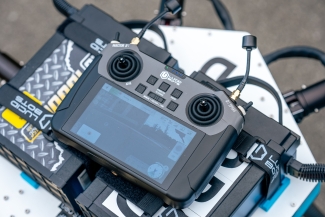 Close-up of a Lucid Bots handheld remote controller with a digital display screen and joysticks, resting on a white surface alongside industrial batteries.