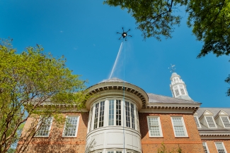 A specialized industrial drone hovers above a multi-story red brick building, spraying a stream of water onto the roof under a clear blue sky.