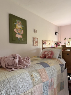 A bed covered in a pastel patchwork quilt and a pink bow-shaped pillow sits next to a wall featuring a large vintage-style illustration of a girl in a flower hat.