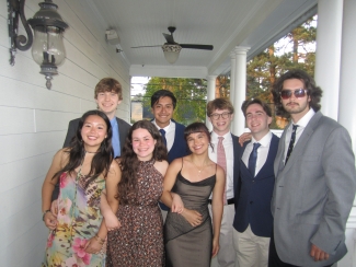 A group of seven friends in formal evening wear poses together on a white porch under a ceiling fan.