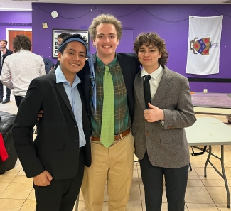 Three young men in suits smile for a photo in a room with purple walls, including one man on the left wearing a blue tie tied around his forehead as a headband.