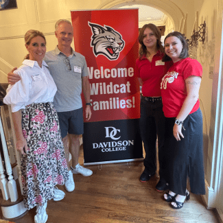 Two men and two women stand on a hardwood floor next to a red and black vertical banner that features a wildcat logo and the text, "Welcome Wildcat Families! Davidson College."