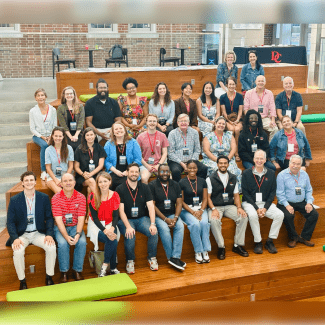 A diverse group of approximately twenty-five people sit on wide, tiered wooden bleachers indoors for a large group portrait.