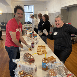 Volunteers wearing gloves smile while standing at a long table covered in loaves of bread and jars of peanut butter and jelly, preparing sandwiches in a large room.