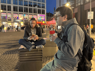 Two people sit on metal benches in a cobblestone city square at dusk, eating street food with illuminated modern buildings in the background.