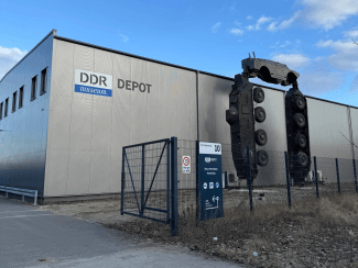 The exterior of the DDR Museum Depot in Berlin, featuring a large corrugated metal building with a sculpture of an armored vehicle perched atop two vertical tanks.