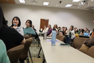 A young woman smiles while speaking into a microphone during a seated presentation in a lecture hall filled with attentive students.