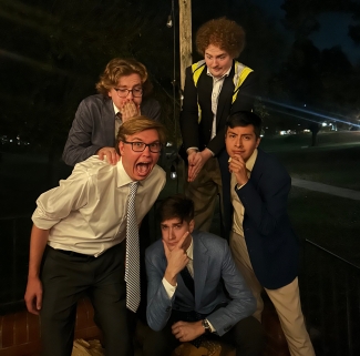 Five young men in business attire make playful, exaggerated facial expressions and poses while gathered outside at night under hanging string lights.