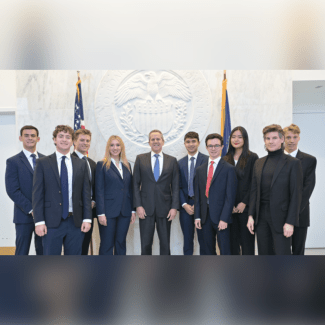 A group of nine young adults in professional business attire and one older man in a suit pose together in front of the large, circular stone seal of the Federal Reserve System and American flags