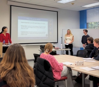 Three students lead a presentation in a classroom titled "Policy Recommendation," displaying bullet points regarding the Federal Funds Rate, balance sheet reductions, and inflation communication to an audience.