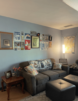 A cozy living room featuring a green sofa adorned with a lighthouse-patterned throw and floral pillows, situated beneath a gallery wall of eclectic art and posters.