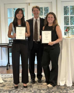 Two students smile and hold "C. Louise Nelson Award" certificates while standing on either side of a faculty member in a dark suit and patterned tie.