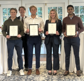 Four students stand in a row holding framed "Rostan Award" certificates, with a faculty member smiling behind them in a room with a patterned rug.