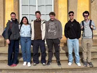 Six young adults stand in a row smiling for a group photo on stone steps in front of a yellow building with large white-framed windows.