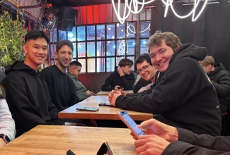 A group of five young men sit around a wooden table in a dimly lit restaurant, smiling at the camera while other patrons are visible in the background.