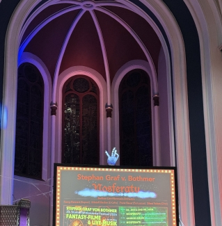 Inside a vaulted building with arched windows, a large screen displays a poster for a "Nosferatu" silent film concert festival by Stephan Graf von Bothmer, topped with a decorative glowing hand sculpture.