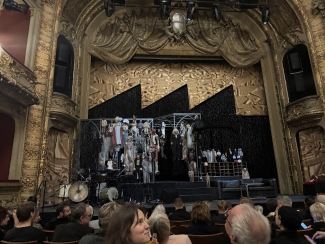 An ornate, gold-detailed theater stage featuring a complex set with numerous hanging puppets and a black sawtooth backdrop, viewed from the perspective of an audience in the darkened seating area.