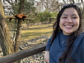 A smiling woman takes a selfie in front of a zoo enclosure where a red panda is resting on a tree branch in the background.
