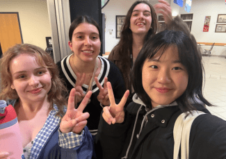 Four young women smile for a close-up selfie in an indoor common area, with two making peace signs at the camera.