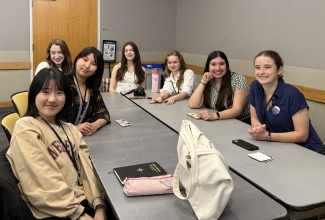 Seven young women sit around a long gray conference table, smiling at the camera during a group session.