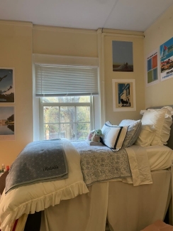 A twin bed with ruffled white and blue floral bedding is positioned under a window, flanked by cream-colored walls decorated with vintage-style art prints.