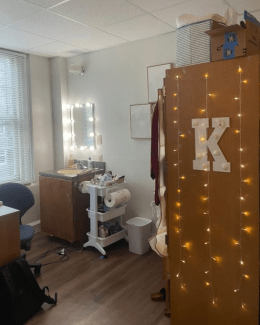 A room featuring a vanity area with a lighted mirror and sink, positioned next to a tall wooden wardrobe decorated with warm string lights and a large white letter "K."