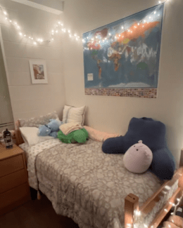 A twin bed with a floral duvet and several plush pillows is positioned beneath a large world map and a strand of warm white string lights.