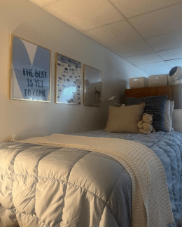 A bed with light blue quilted bedding and a teddy bear is set against a wall featuring three framed prints, including one that reads, "The best is yet to come."
