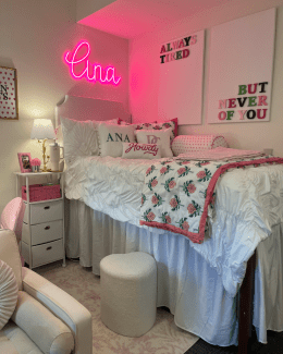 A highly decorated dorm room featuring a lofted bed with white ruffled bedding, a bright pink "Ana" neon sign, and wall art reading "Always Tired But Never Of You."