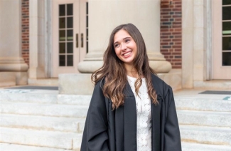 Courtney wears a black graduation gown over a white lace dress and smiles while looking off-camera on brick college steps.