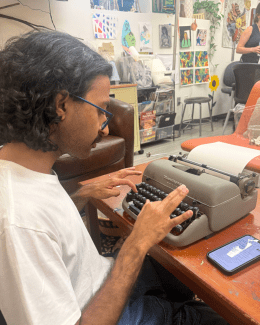 A student with curly hair and glasses types on a vintage Remington typewriter at a wooden desk in a room filled with colorful art.