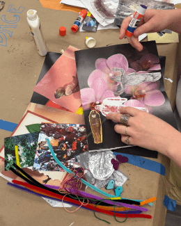 Close-up of hands assembling a collage on brown paper using glue sticks, magazine cutouts of flowers and figures, and colorful pipe cleaners.