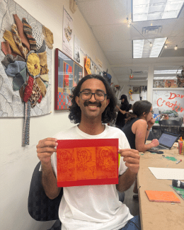 A smiling student with glasses and curly hair holds up a red piece of paper featuring three orange linocut prints of a face while sitting in a brightly decorated art studio.