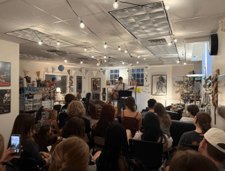 An audience of students watches a live musical performance by a guitarist under warm string lights in a decorated community art space.
