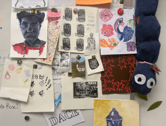 A whiteboard is covered in a variety of student artworks, including a portrait, fish sketches, and a long blue knit caterpillar plush.