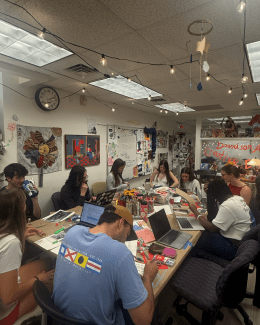 A group of students sits around a large, cluttered table in the Davidson Arts and Creative Engagement (DACE) Studio, working on various art projects and using laptops under warm string lights and a whiteboard filled with sketches.