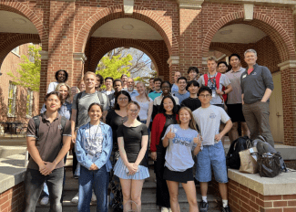 A large, diverse group of students and faculty pose together for a commemorative photo on the steps of a brick building with prominent arched entryways.