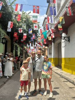 A family of four smiles while standing together on a cobblestone street decorated with numerous international flags strung overhead.