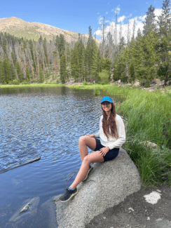 A smiling woman with long brown hair, wearing a white sweater, dark shorts, and a blue cap, sits on a large boulder next to a calm lake with a forested mountain in the background.