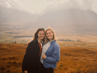Two smiling women stand close together outdoors against a hazy, sweeping landscape of rolling hills and brown fields.